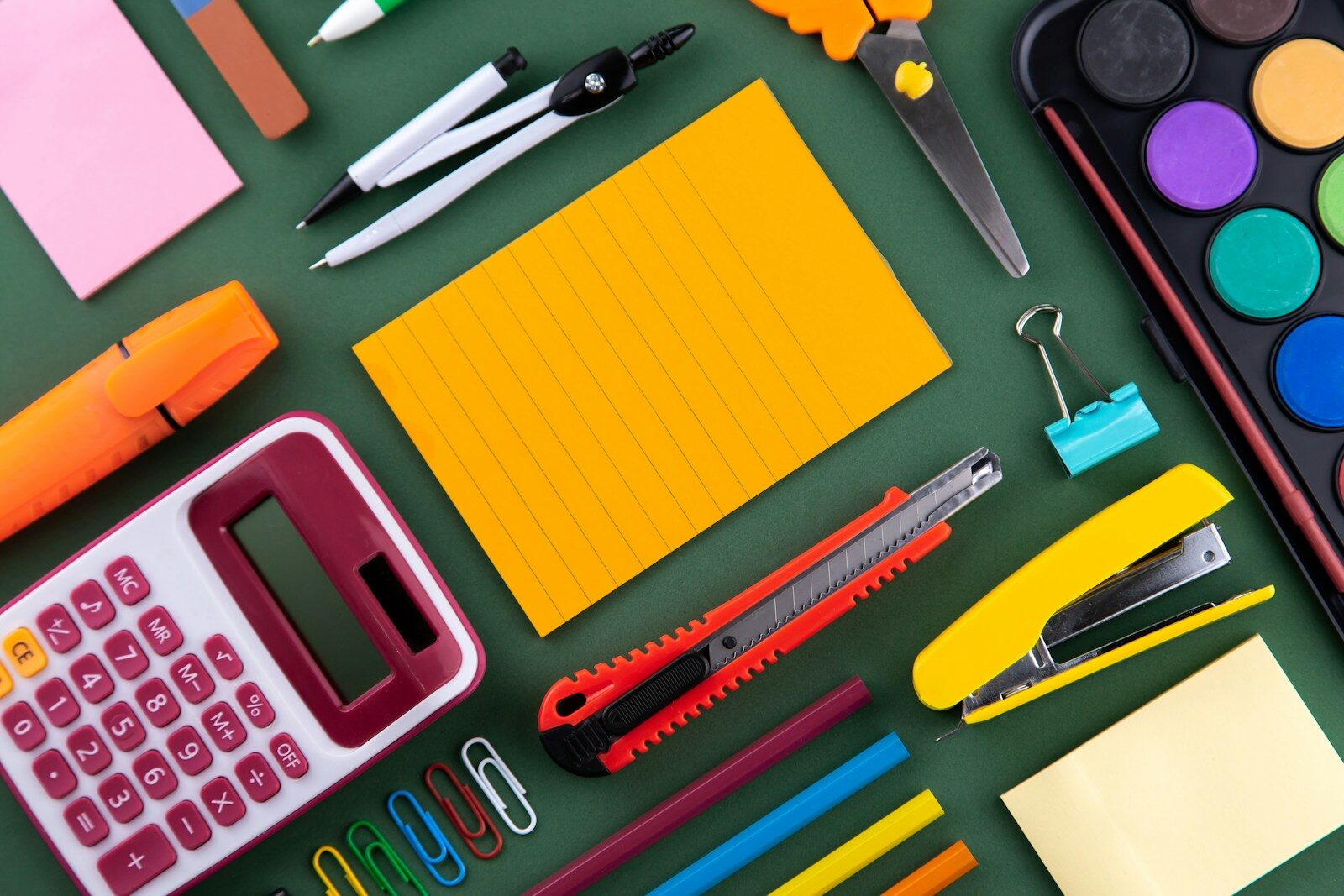 Stationery including a calculator, scissors, stapler, and notepad on a green surface